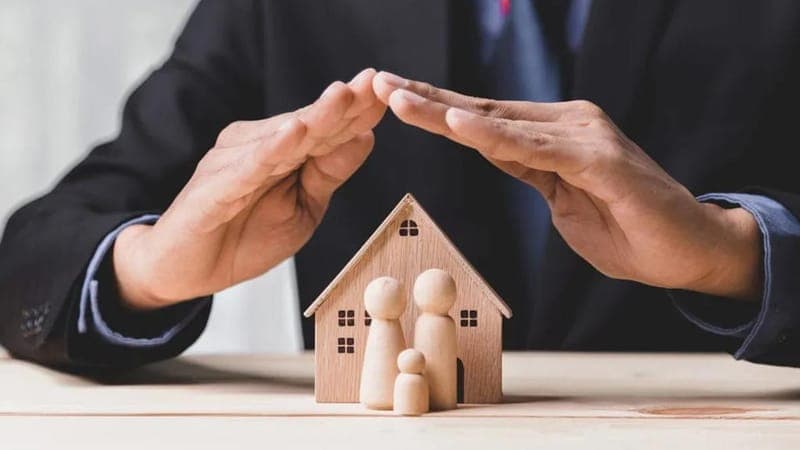 Mãos formando um telhado sobre uma casa de madeira com três figuras dentro